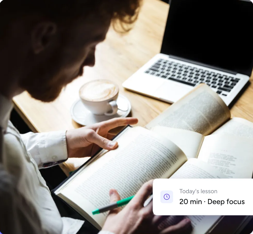 A man reading an open book beside a laptop and a coffee